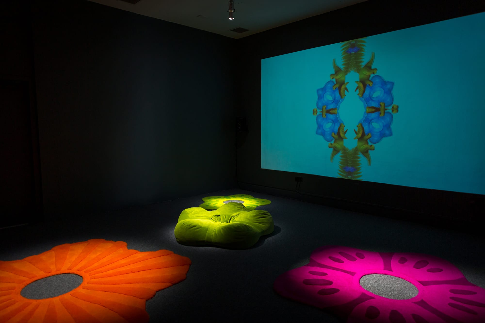 An exhibition hall black walls and a floor. There's a projection on the wall, and four installations (orange, pink, two green) for children to interact with.