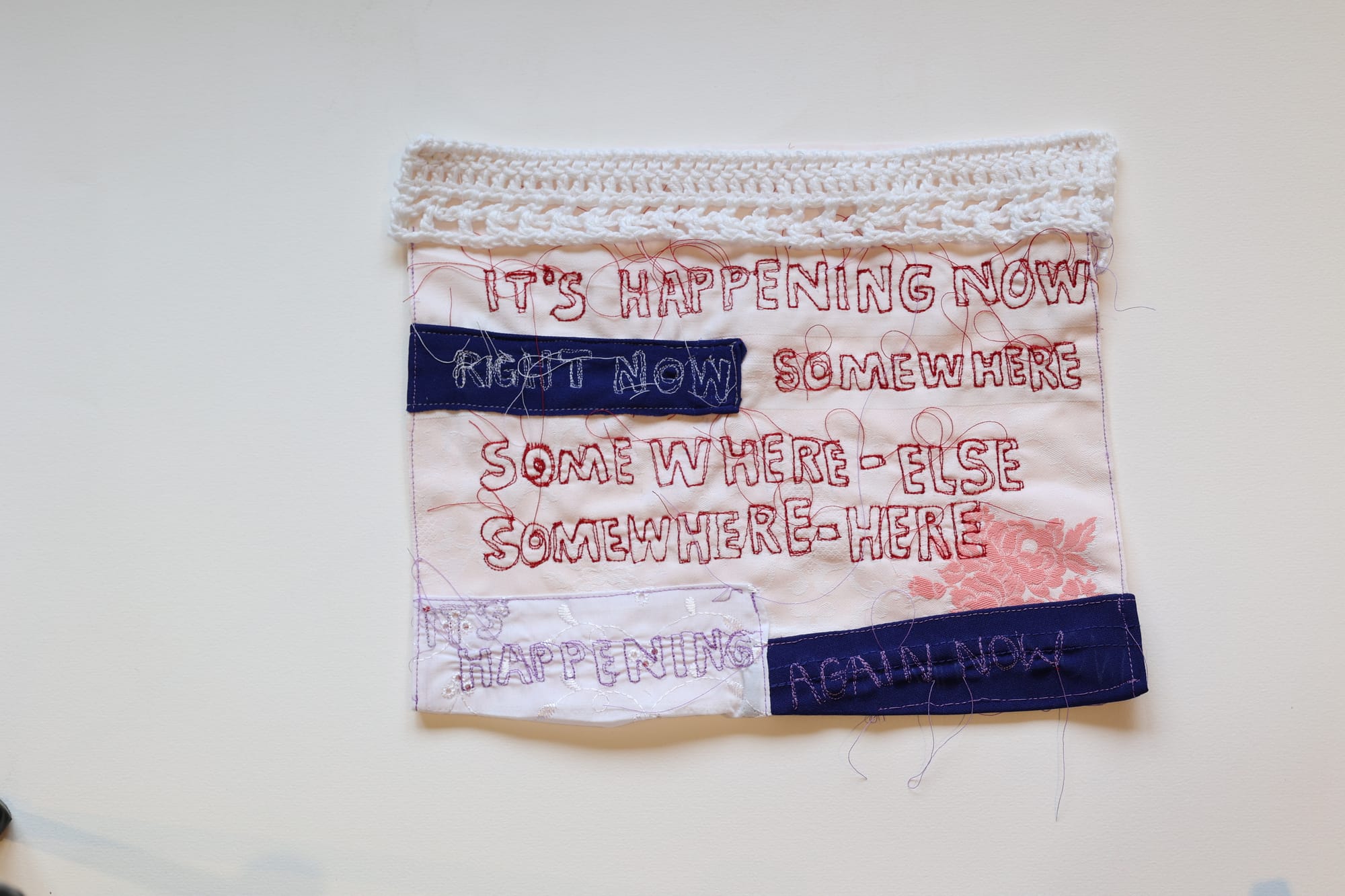A photograph of a piece of Christina Bennett's work. It's a textile with the words in all caps: It's Happening Now / Right Now / Somewhere / Somewhere - Else / Somewhere - Here / Happening / Again Now