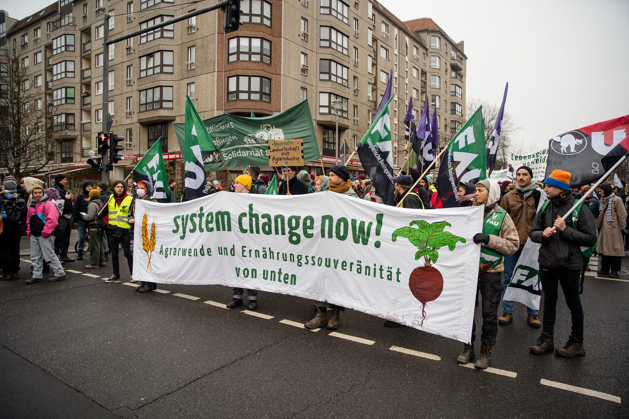 Protestors hold up a white banner saying "system change now" with other signs and flags in the background.