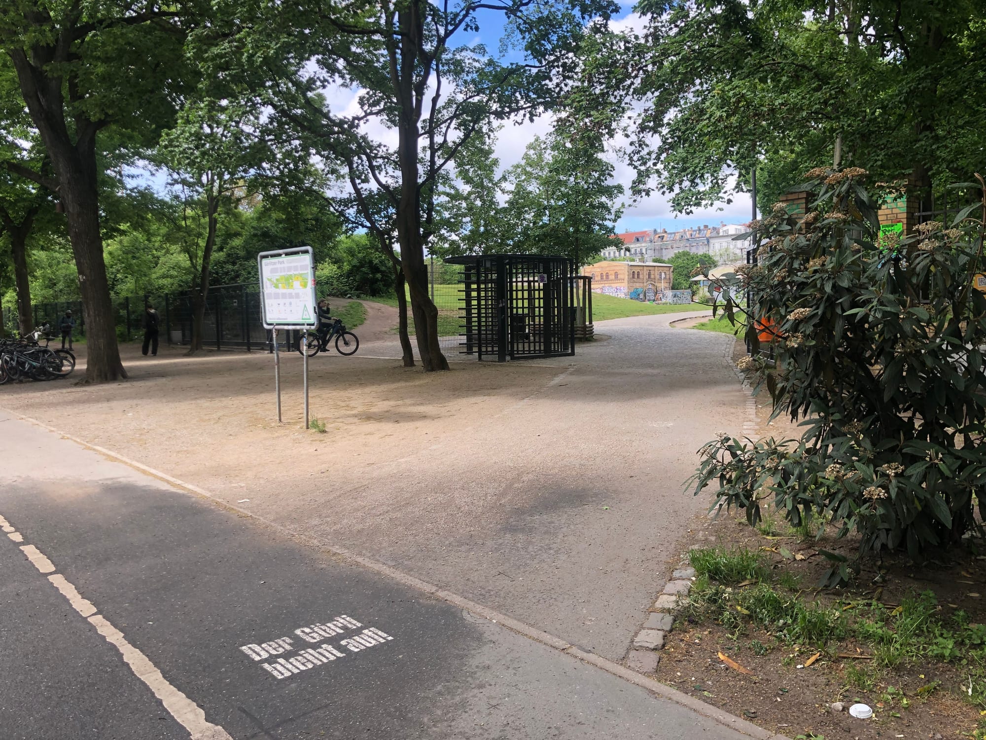 An entrance to a park with a gate. There are many trees and you can see buildings in the distance through them. There's a cycle path with "Der Görli bleibt auf!" stenciled on.