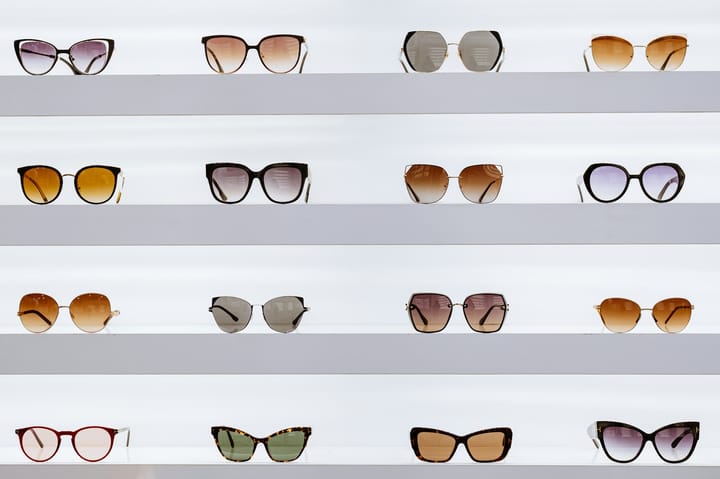 Different pairs of glasses, four rows and four columns, are arranged on white shelves against a white wall. Looks like a glasses store display.