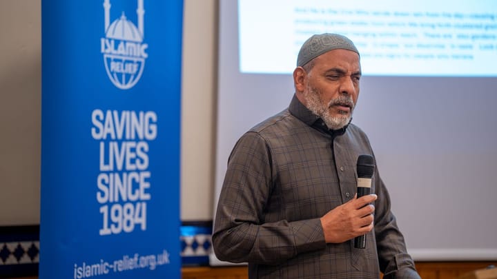 A Muslim man is holding a microphone. Behind him is a blue banner with the Islamic Relief logo, as well as a whiteboard with blurred text projected onto it.