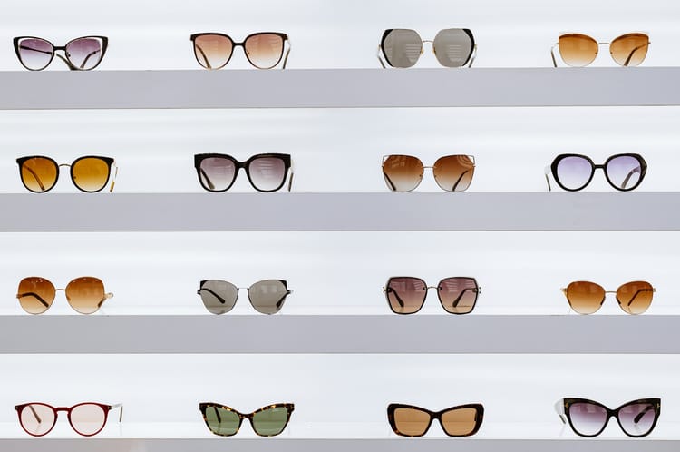 Different pairs of glasses, four rows and four columns, are arranged on white shelves against a white wall. Looks like a glasses store display.