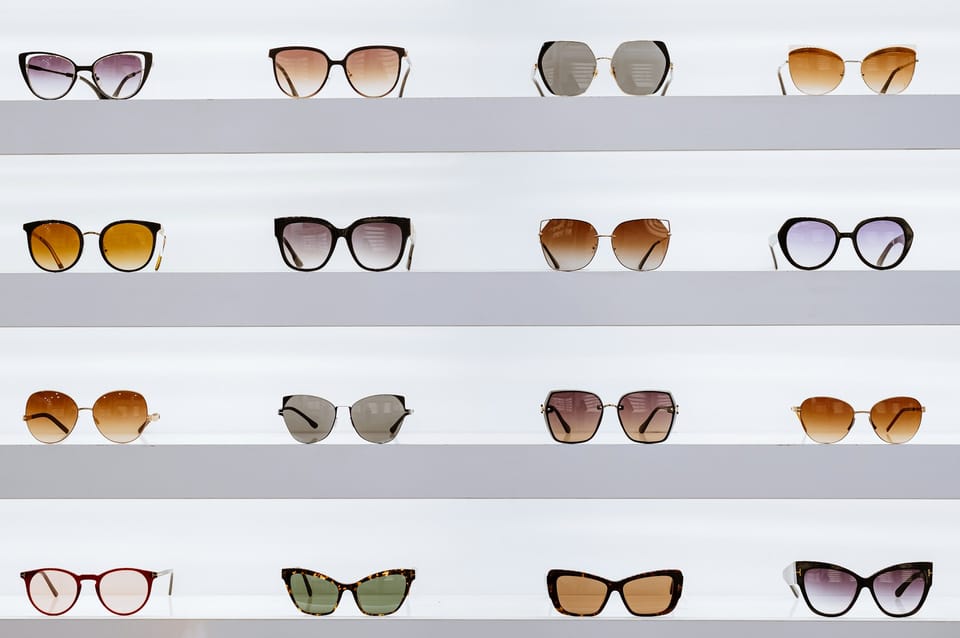 Different pairs of glasses, four rows and four columns, are arranged on white shelves against a white wall. Looks like a glasses store display.